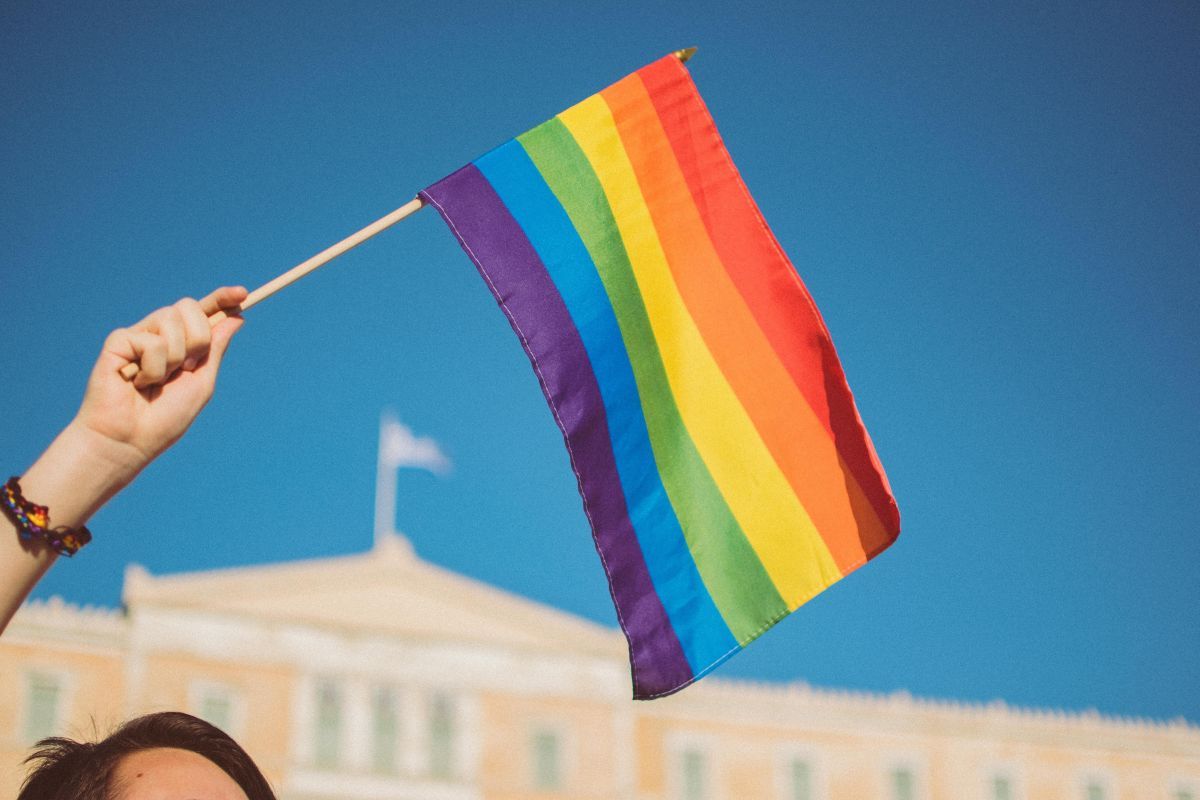 A person holding the iconic rainbow-coloured LGBTQ+ flag with the Greek Parliament building in Syntagma, Athens, in the background. Photo by Stavrialena Gontzou on Unsplash.